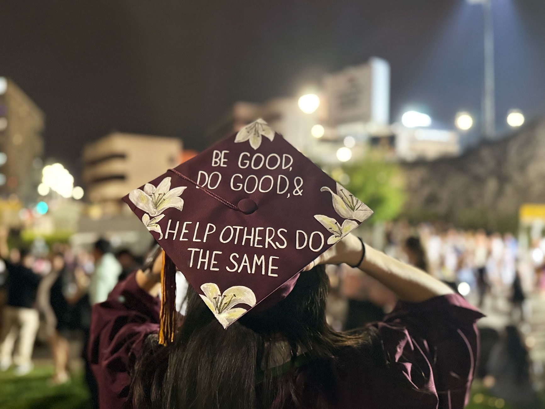 Morgan Kubasko's hand-painted graduation mortarboard, photographed from behind on a windy night at ASU's Sun Devil Stadium, May 2025. The cap is maroon with white-painted lilies and reads, in white block letters: 'BE GOOD, DO GOOD, & HELP OTHERS DO THE SAME.'