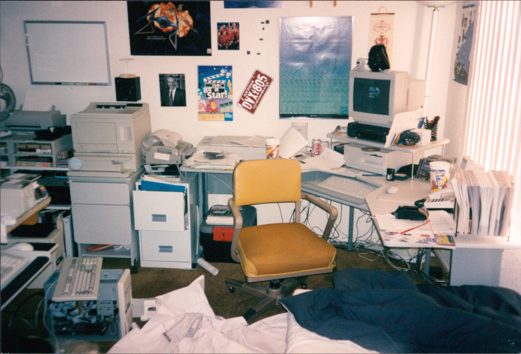 A photograph of Rob Kubasko's actual office in 1996. A corner desk holds a beige-cased Mac and a thermal-roll fax machine. Behind it, the wall is pinned with a Bob Dole campaign headshot, a Star Trek poster, an Arizona license plate reading 'DVX 805', and a printed sign that says 'Be a Star.' On the floor: a yellow office chair, file cabinets, and a half-disassembled tower computer.