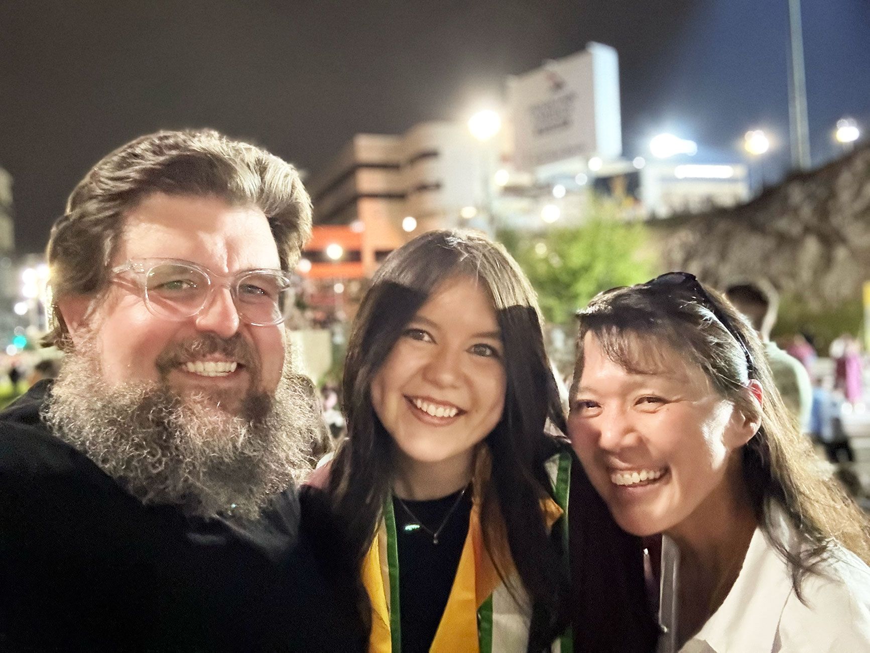 A late-night family selfie at ASU's graduation grounds in May 2025: Rob Kubasko on the left in glasses with a full grey-brown beard, Morgan Kubasko in the middle wearing her maroon graduation gown and gold honors stole, and her mother Margie on the right with sunglasses pushed up on her head. All three are smiling; the lit stadium glows behind them.